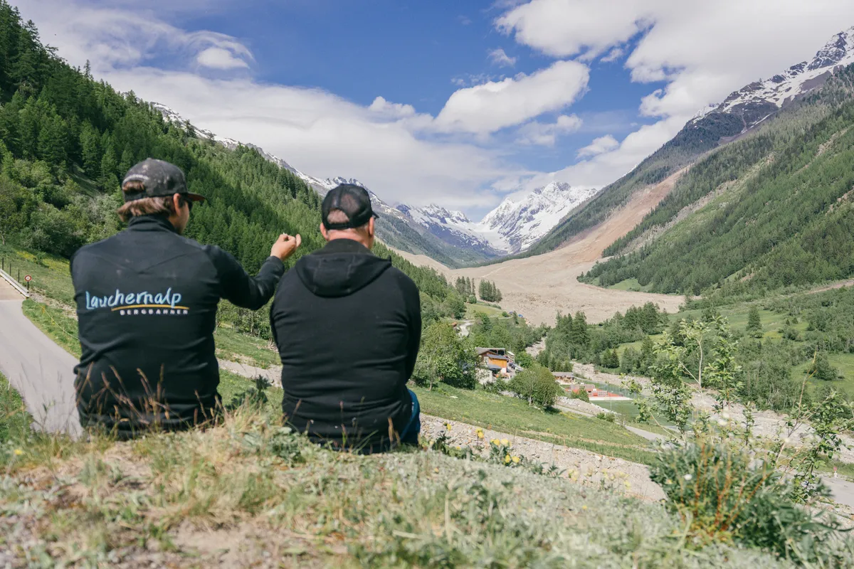 Zwei Männer von hinten auf einem Wiesenhang zeigen ins Tal auf einen Gletscher zwischen den Bergflanken