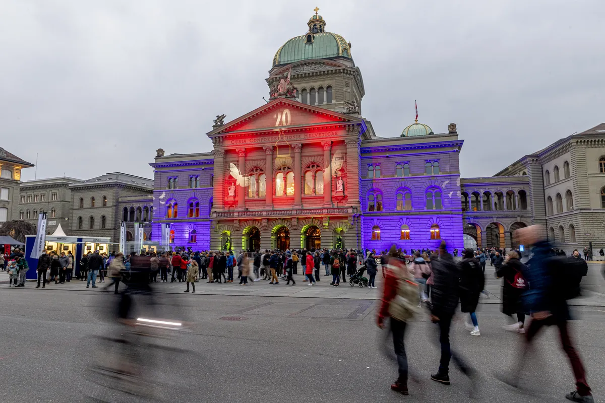 Bundeshaus in Bern farbig illuminiert, davor Publikum und vorbeiziehende Passanten